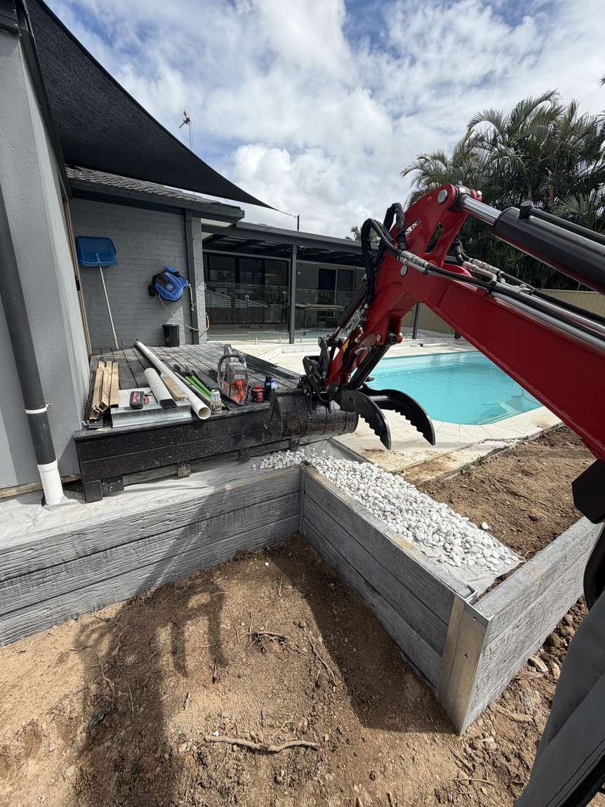 The excavator arm working over a retaining wall corner beside a pool